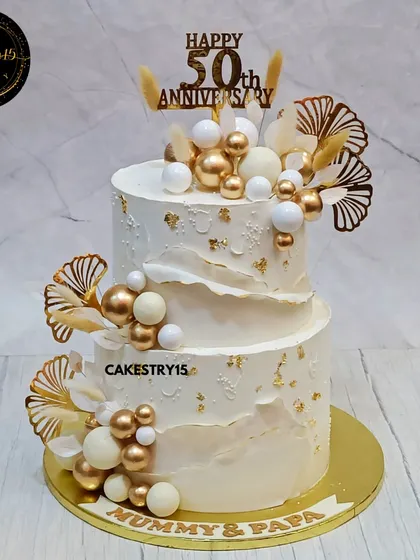 An elegant 5kg, two-tier chocolate cake for a 50th anniversary. The ivory-colored cake is decorated with white and gold decorative balls, golden ferns, and a "Mummy & Papa" nameplate.