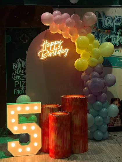A colorful fifth birthday setup with a large light-up marquee number '5'. The decor includes a rainbow balloon garland and a neon 'Happy Birthday' sign.