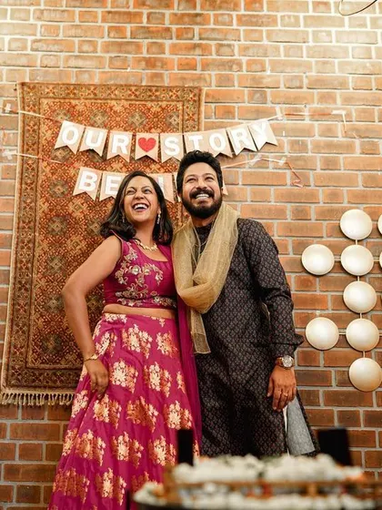 The couple stands together, smiling, at their engagement party with a "Our Story Begins" banner in the background.
