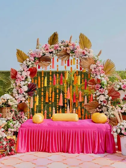 A beautiful Mehendi setup with a pink and gold theme, featuring a floral arch, traditional tassels, and elegant seating for the bride.