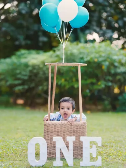 An adorable shot of a one-year-old peeking out of a hot-air balloon basket during his outdoor birthday shoot. The 'ONE' sign clearly marks the special occasion.