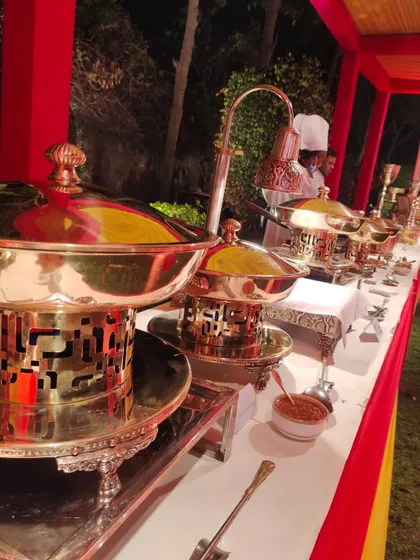 A close-up of the traditional brass and copper serving dishes, known as 'chafing dishes' or 'bain-maries', keeping the food warm. This setup is part of a wedding catering service in a colorful outdoor tent.