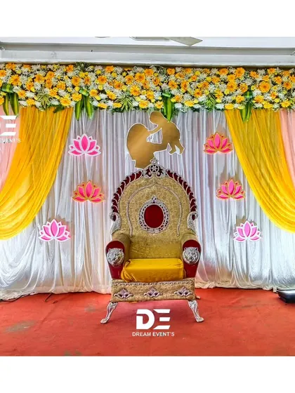 A traditional baby shower stage featuring a beautiful throne chair and a backdrop of yellow and pink drapes. The lotus flower cutouts and mother-child silhouette add a meaningful touch.