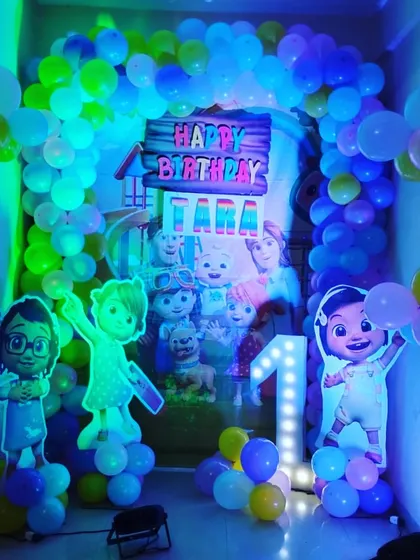 A Cocomelon-themed first birthday party decoration for a home. The setup includes a balloon arch, character cutouts, and a light-up number '1'.