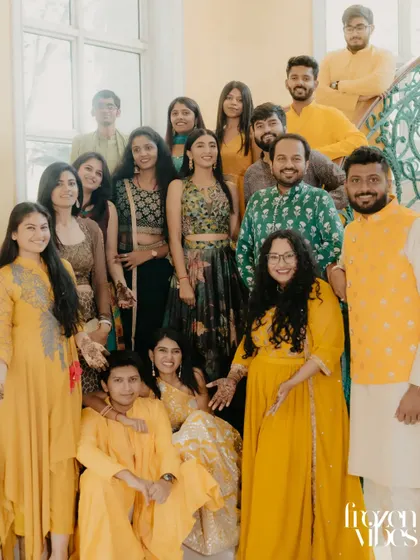 A large group of friends and family in vibrant yellow and green traditional wear pose together on a staircase. This photo captures the festive atmosphere and collective joy of a Haldi or Mehendi ceremony.