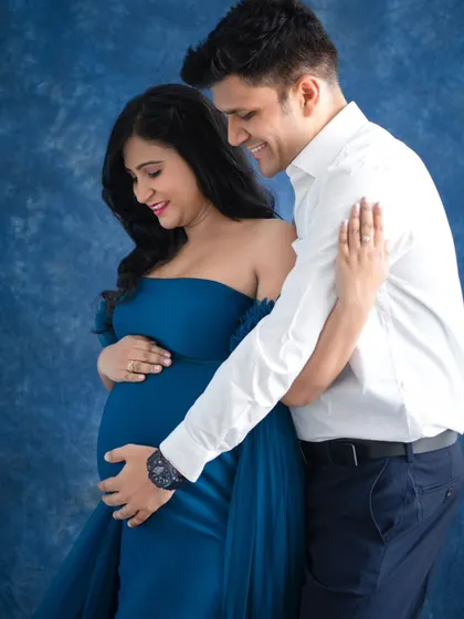 The husband gently embraces his wife from behind, both of them looking down at the baby bump with soft smiles. It's a portrait of quiet, shared joy.