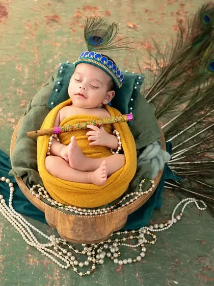 A sleeping newborn styled as Krishna, holding a flute and nestled in a basket with a beautiful peacock feather arrangement.