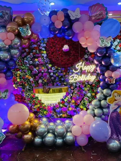 A grand Disney Princess themed first birthday party. The setup features a flower wall, balloon arrangements, cutouts of Ariel and Rapunzel, and a "Happy Birthday" neon sign.