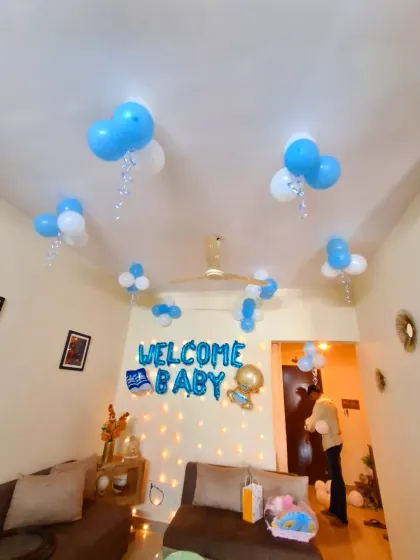 A living room decorated to welcome a new baby. The setup includes a 'Welcome Baby' foil banner, a fairy light wall, and helium balloons on the ceiling.