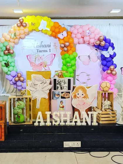This fairy-themed first birthday setup is full of playful details. It includes fairy cutouts, rainbow-colored balloons, and wooden crates that add a rustic, enchanted forest feel.