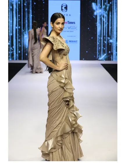 A side view of the beige draped saree gown on the runway. This angle showcases the cascading ruffles and the expertly tailored fit of the corset bodice.