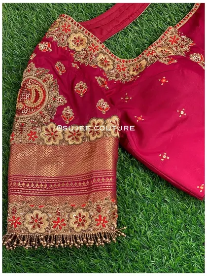 A custom maroon bridal blouse with a sweetheart neckline. The sleeve features a personalized touch with an embroidered initial 'D' inside a traditional motif, surrounded by floral zardozi work.