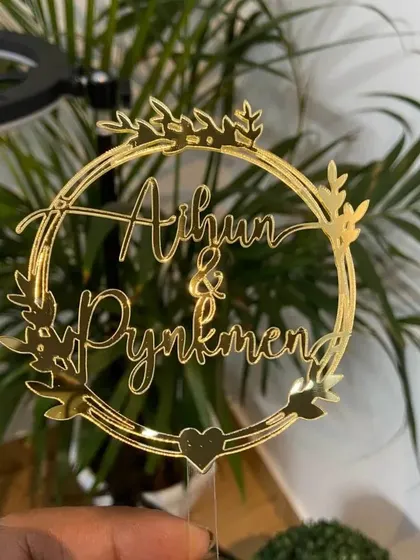 The "Aihun & Pynkmen" wreath topper held up, showing its intricate details and beautiful gold finish.