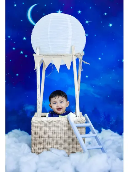 A whimsical hot air balloon themed photoshoot. A smiling toddler sits in a basket prop, seemingly floating among the clouds against a starry night backdrop.