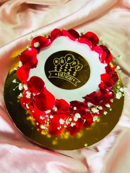 A simple and elegant birthday cake topped with a halo of fresh red rose petals and baby's breath. The golden "Happy Birthday" plaque in the center adds a celebratory touch.