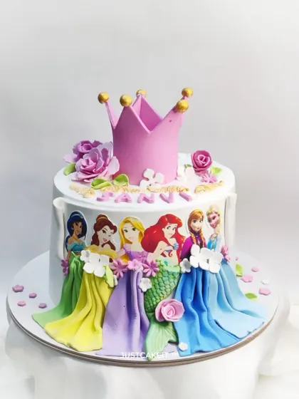 A simple and elegant Disney princess cake with a large, edible crown on top. The design features the princesses' dresses as a beautiful, draped border.