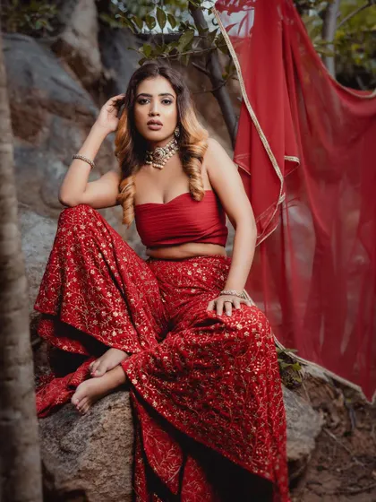 A full view of the model seated in the red sharara set, showing the wide-legged pants and bandeau-style top.