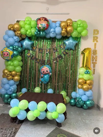 A Cocomelon birthday setup for a first birthday, complete with a balloon arch, shimmer backdrop, and a balloon pillar with the number '1'. The child's name is displayed with foil letter balloons.