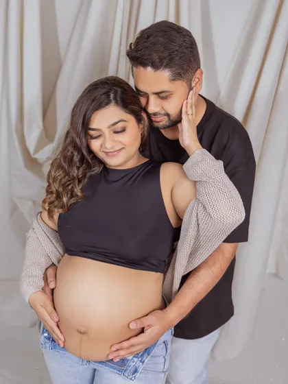 A close-up showing the love and connection. The father-to-be embraces his partner from behind, both looking down at her bump with gentle smiles, capturing a quiet and loving moment.