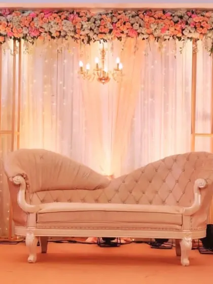 An elegant chaise lounge sits before a backdrop of sheer curtains and a crystal chandelier, framed by a delicate arrangement of peach and white flowers.