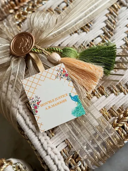 Detail of the personalized gift tag for the engagement invitation hamper. The tag features a delicate peacock illustration and is attached with a colorful tassel, echoing the main wedding theme.