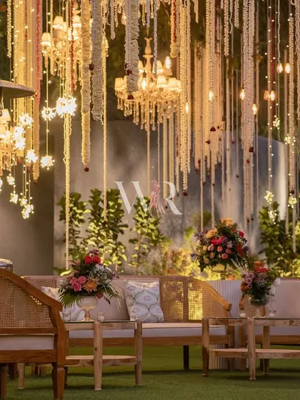 A cozy lounge area set up for a wedding ceremony, under a canopy of cascading mogra and lit by the warm glow of chandeliers. The natural wood furniture adds a touch of rustic elegance to the traditional setting.