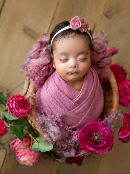 A sweet baby girl nestled in a basket, surrounded by vibrant pink roses. The wood background adds a rustic, warm touch to this floral newborn portrait.