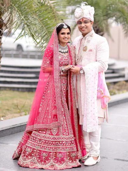 A classic, full-length wedding portrait of the couple posing for the camera, showcasing the intricate embroidery on the bride's bright pink lehenga.