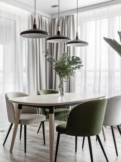 A modern dining setup with mismatched chairs in green and grey tones, set against a backdrop of sheer curtains. The pendant lights add an industrial-chic touch.