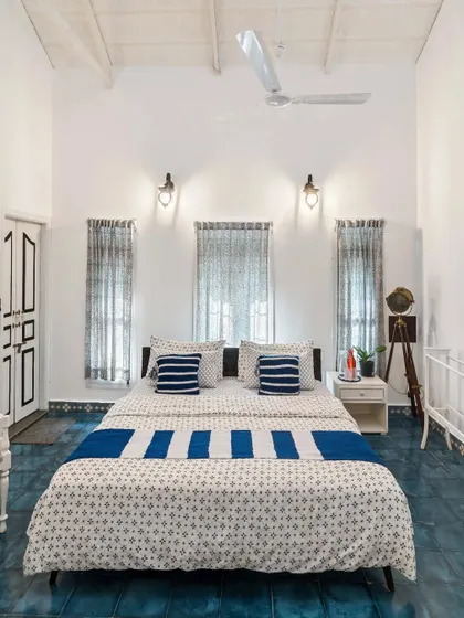 A bright and airy bedroom in a Chikmagalur homestay, with a high vaulted ceiling and blue tiled floors. The simple, clean aesthetic is accented by blue and white block-printed textiles.
