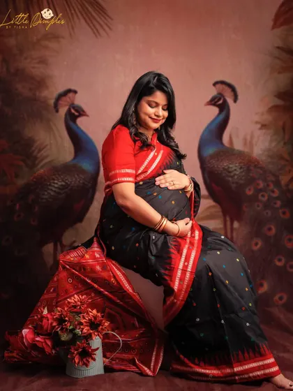 A beautiful fine art portrait of a mom-to-be in a traditional black and red saree, seated with peacocks in the background. The colors and composition are just stunning.