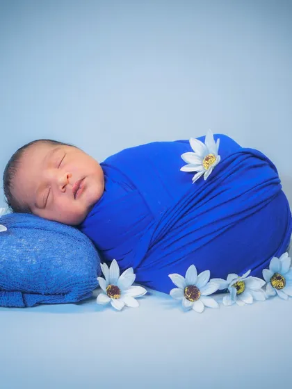 A perfectly swaddled newborn in a vibrant blue wrap, surrounded by delicate white flowers. This simple "potato sack" pose is safe, comfortable, and always adorable.
