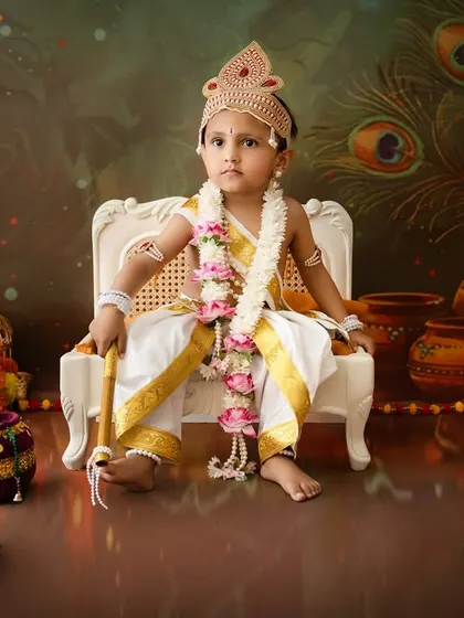 A regal portrait of a toddler dressed as Krishna, sitting on a white chair against a peacock feather backdrop.