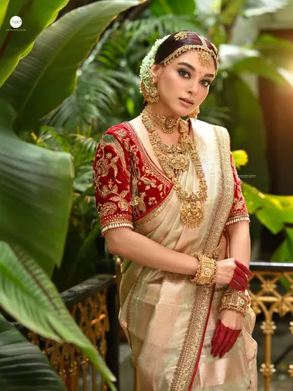 A full-length bridal portrait featuring a stunning off-white and red saree. The lush green background makes the traditional attire pop, and the use of natural light adds a soft, radiant glow to the scene.