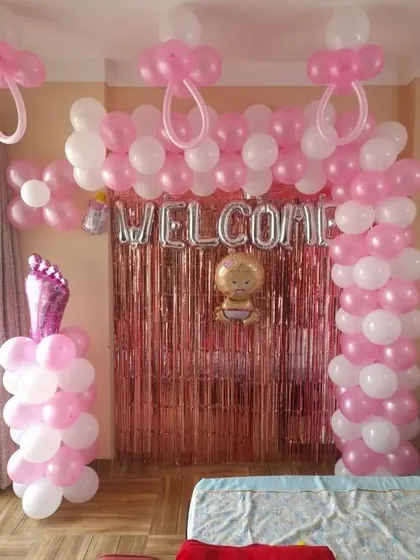A simple and sweet "Welcome" decoration for a newborn. This setup includes a pink and white balloon arch, a foil fringe curtain, and baby-themed foil balloons, perfect for a home celebration.