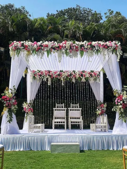 This is an elegant vedi mandap I designed for an outdoor day wedding. The structure is draped in clean white fabric, with lush floral arrangements of pink and white roses on top and along the pillars, creating a modern yet timeless look for the pheras.