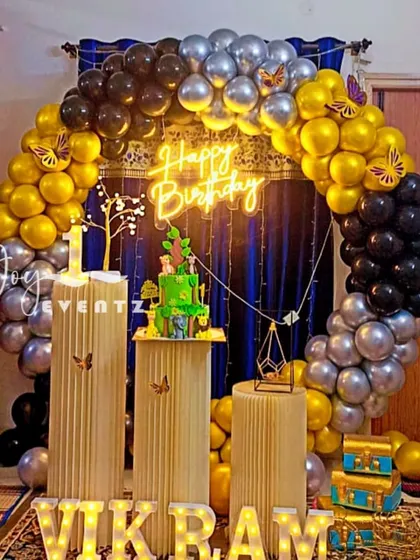 I created this elegant balloon ring decoration for Vikram's first birthday. The arch of gold, black, and silver balloons is set against a dark curtain, with fluted plinths for the cake and light-up marquee letters.