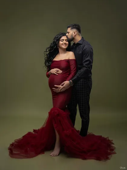 A tender moment in the studio. He gently kisses her forehead as she cradles her bump, both dressed in elegant, coordinating outfits.