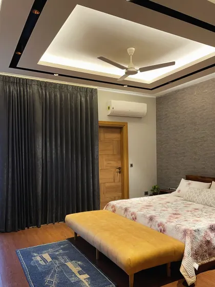 This view of the grey and mustard bedroom shows the layered lighting design, with a modern false ceiling and ambient cove lighting. The dark grey curtains provide a strong contrast to the lighter walls.