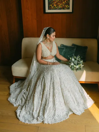 A bride seated on a couch, bathed in warm sunlight. Her intricate white lehenga and relaxed pose create a portrait of modern bridal elegance.