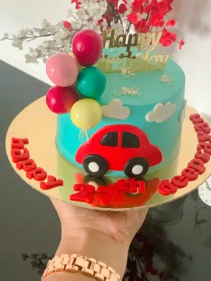 A bright and happy car themed cake. This small cake features a red car, fondant clouds, and a bunch of colorful balloons.