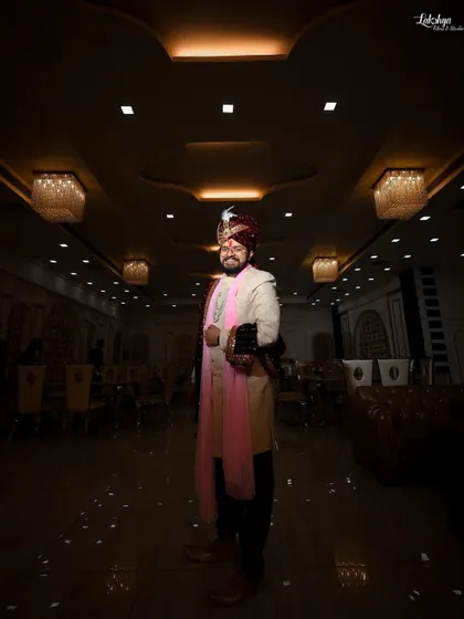 A dramatic, wide shot of the groom standing alone in the empty banquet hall. The lighting creates a sense of anticipation and puts him in the spotlight.