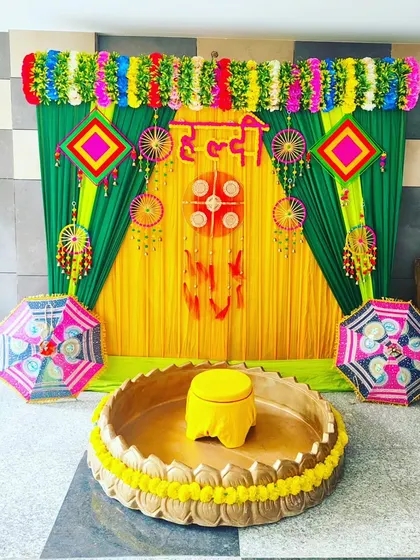 This is a classic Haldi ceremony setup. It features a bright yellow and green backdrop, a traditional brass urli for the ritual, and decorative umbrellas, creating a perfect, joyful setting for your function.