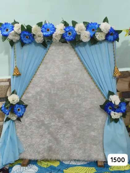 Blue drapes frame a white textured panel with blue and white artificial flowers, advertised at ₹1,500 for ready-made Ganpati decor.
