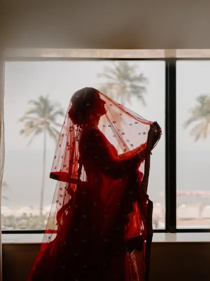 A silhouette of the bride against a window, with palm trees in the background. The red of her lehenga and veil creates a stunning, artistic contrast.