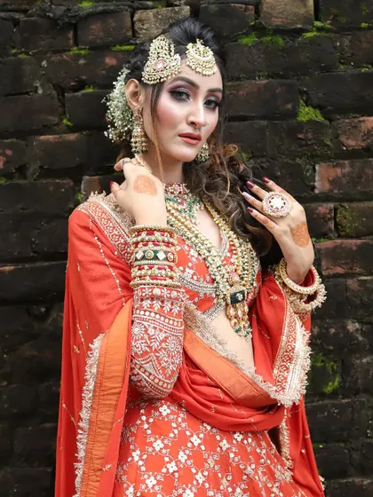 A stunning outdoor shot of a bride in an orange lehenga. Her makeup features dramatic eyes and is paired with a messy, romantic hairstyle.