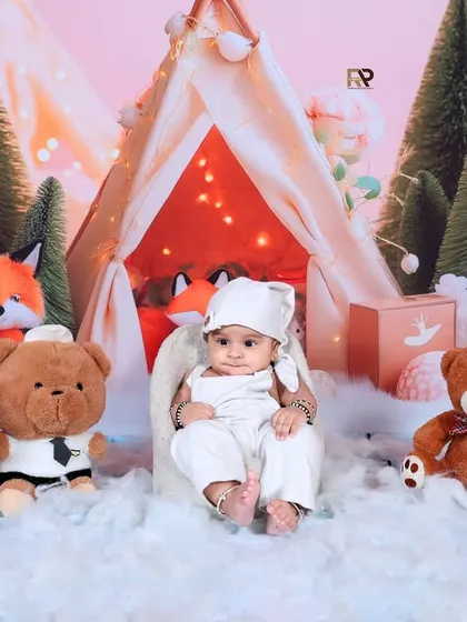 A full view of the winter woodland setup, showcasing the baby surrounded by friendly stuffed animals and pine trees. The soft "snow" and warm lights create a perfect holiday-card photo.