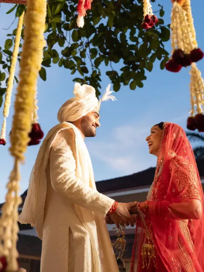 The couple shares a laugh under a canopy of hanging tuberose and marigold garlands. This highlights the blend of traditional floral decor with the natural beauty of an outdoor setting.