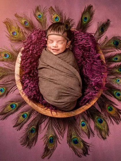 An artistic and elegant portrait, this overhead shot shows a newborn swaddled peacefully in a wooden bowl, encircled by a stunning halo of peacock feathers. The rich colors and textures create a breathtaking piece of art.
