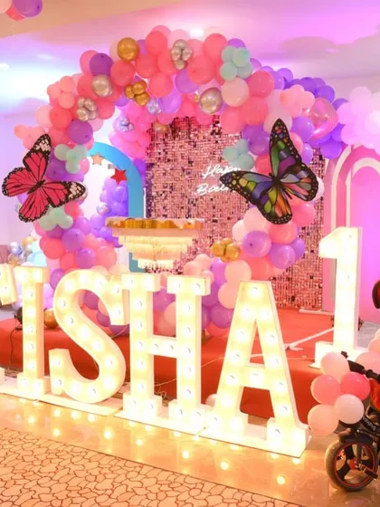 A first birthday party setup with a butterfly theme. The decor includes large marquee letters, a pink sequin backdrop, and a tricycle for the birthday girl.
